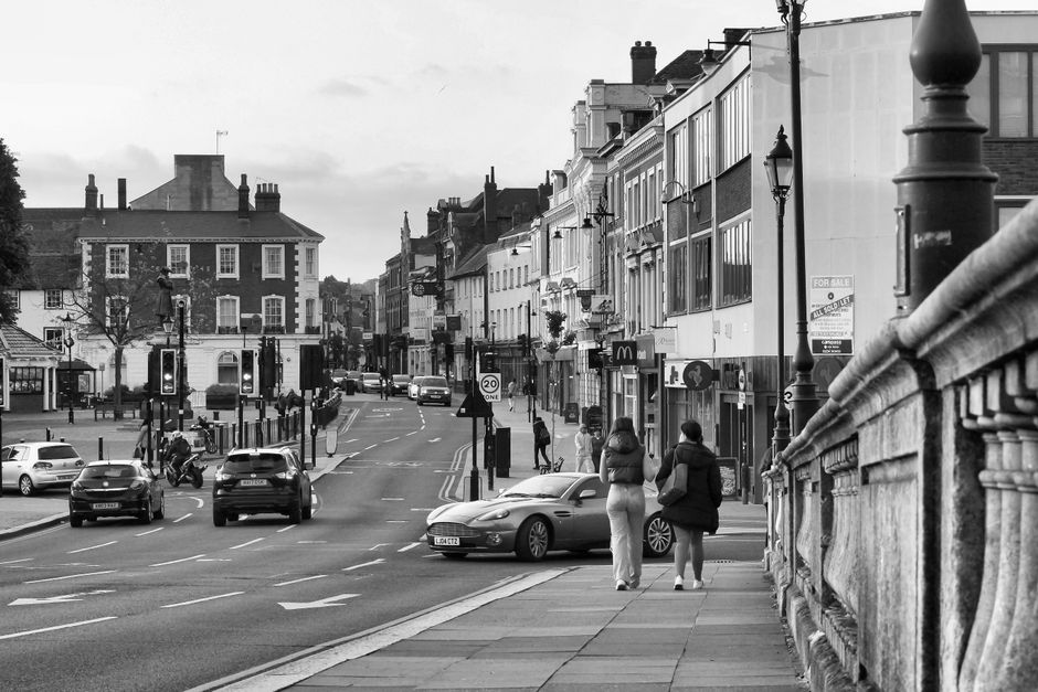 A black-and-white street scene on Marlow High Street featuring pedestrians walking along the pavement, with two women in the foreground engaged in conversation. The street is lined with various shops