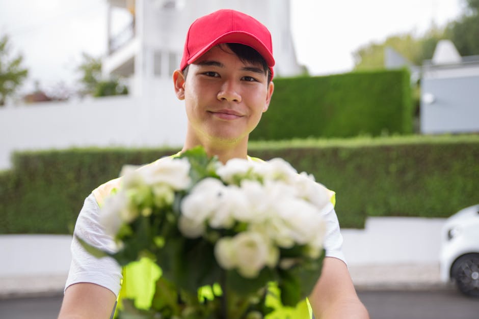 A young male flower delivery courier wearing a red cap and bright yellow high-visibility vest presenting a bouquet of fresh white flowers with green leaves. The bouquet features tightly arranged bloss