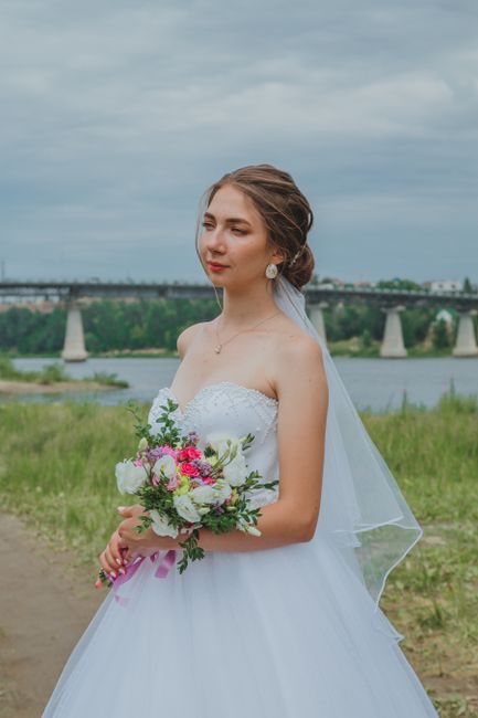 A young woman dressed in a white strapless wedding gown with lace detailing on the bodice, holding a fresh floral bouquet containing pink, white, and purple flowers with greenery accents. She is stand