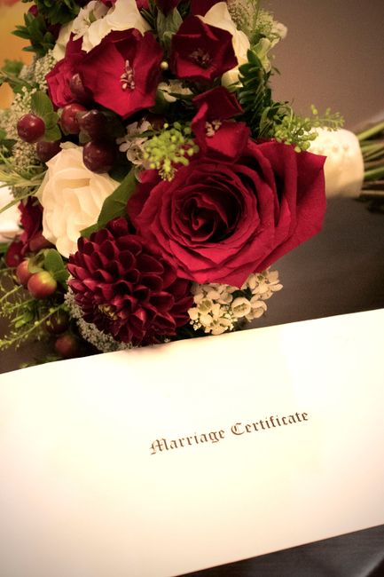A close-up of a floral arrangement featuring deep red roses, burgundy dahlias, white roses, and small green filler flowers, all elegantly arranged with lush green foliage and wrapped partially in tran